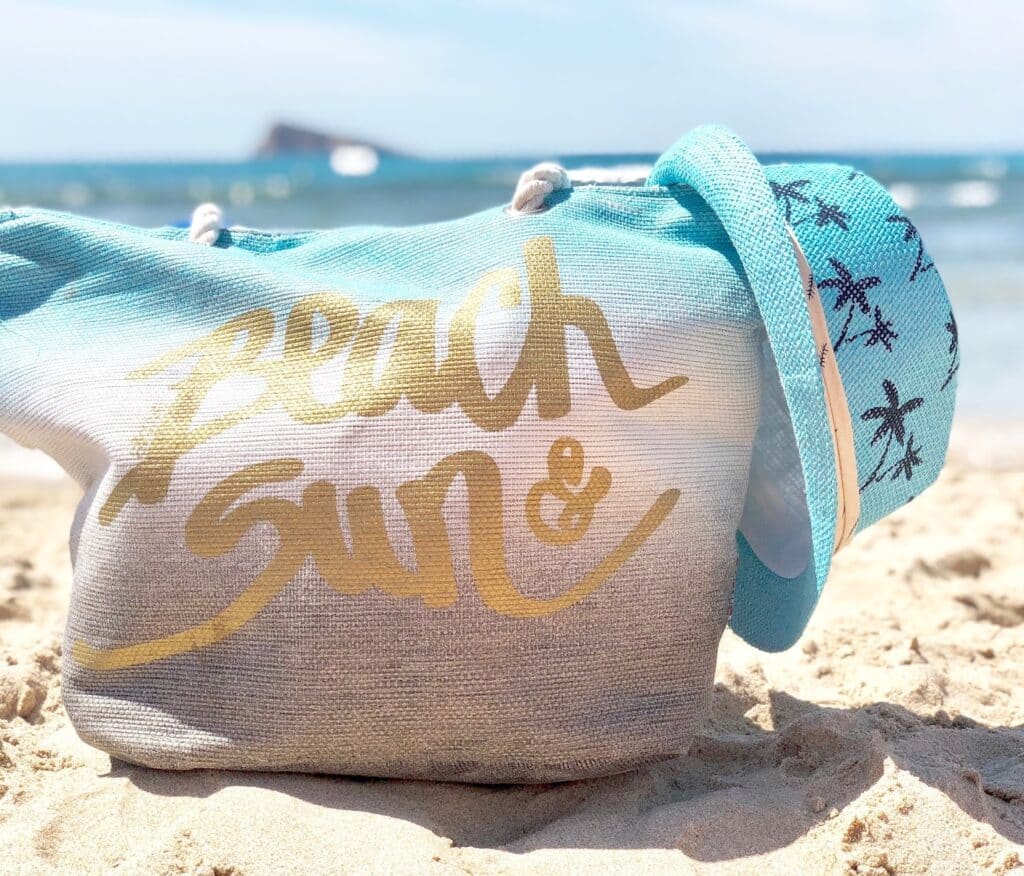 A blue beach bag with "Beach & Sun" in gold sits on the sandy shore beside a turquoise sunhat decorated with starfish, capturing the joy of a family holiday abroad with the sea and sky in the background.