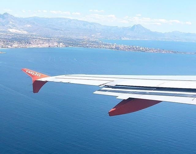 View from an aeroplane window with the easyJet wing above a vast blue sea—coastline, city, and mountains in the distance under a clear sky. A perfect glimpse of a family holiday abroad or your first family holiday adventure together.