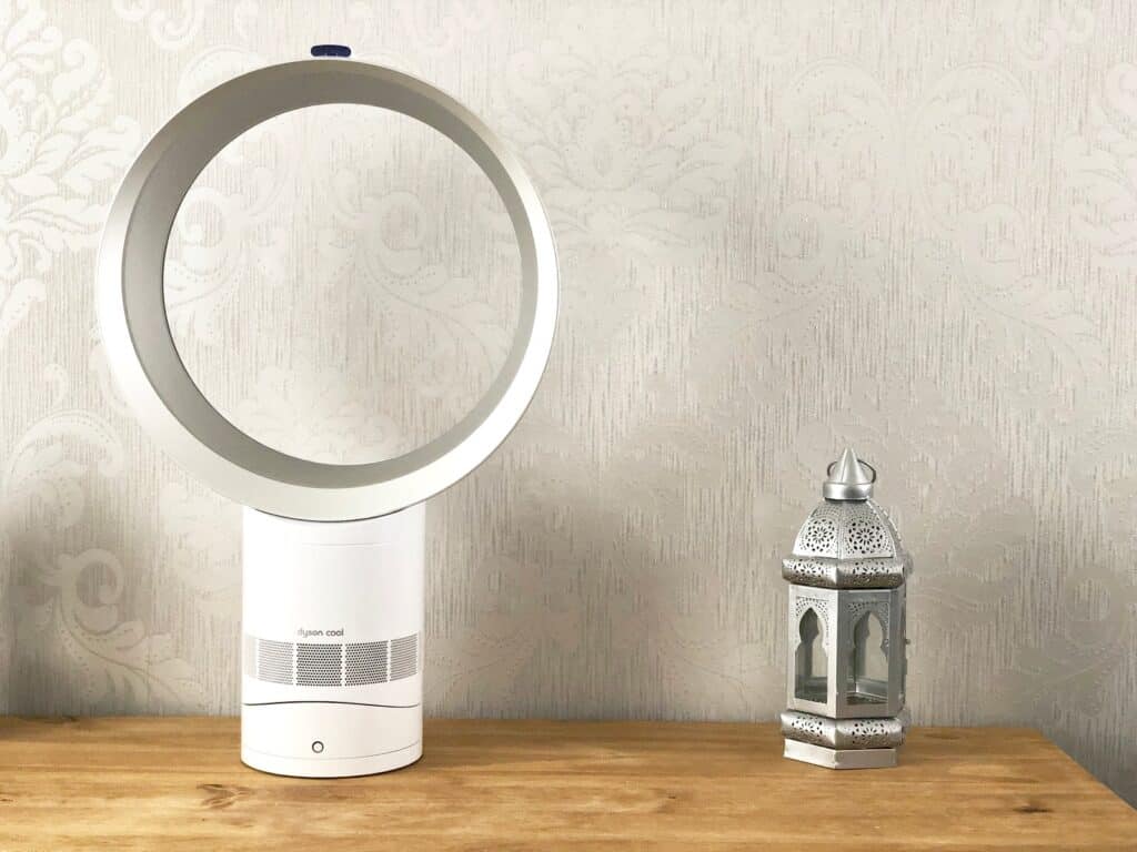 A Dyson AM06 Cool Fan and a small decorative lantern sit on a wooden surface against a light-patterned wallpaper background.