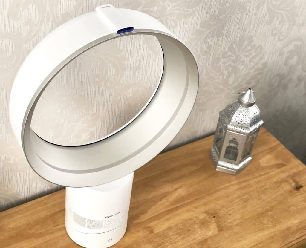 A bladeless white Dyson AM06 Cool Fan sits on a wooden surface next to a decorative silver lantern, with patterned light-coloured wallpaper in the background.