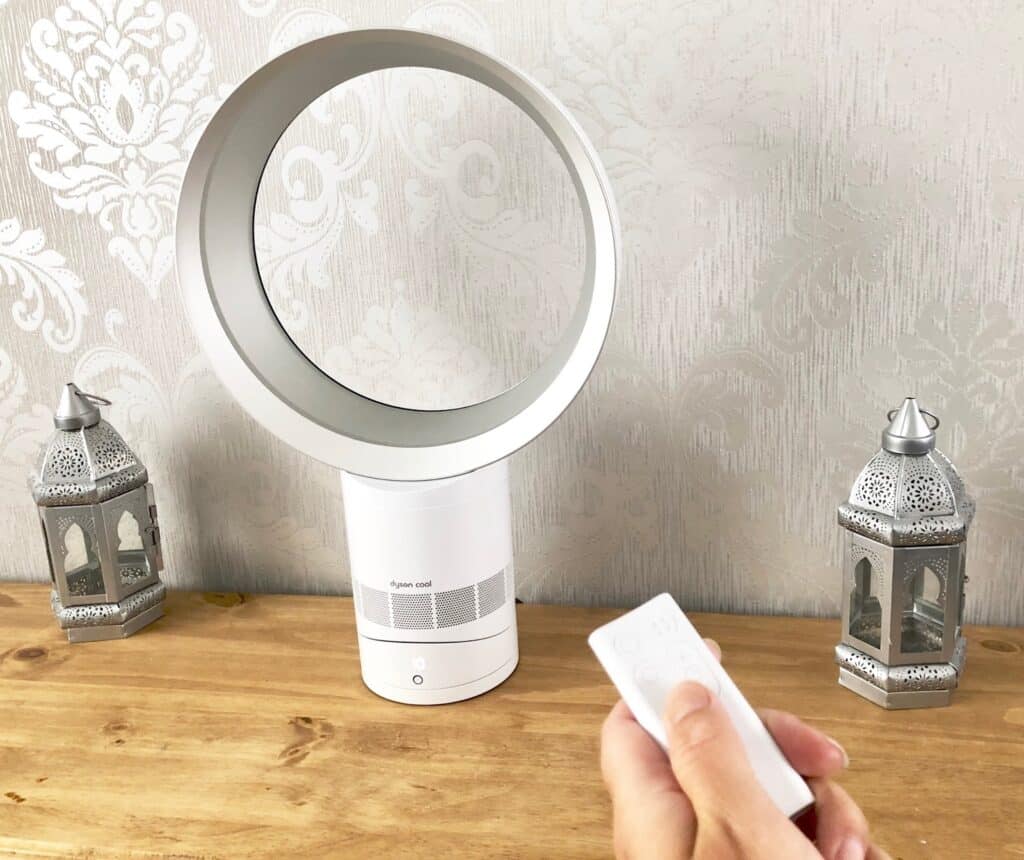 A bladeless white Dyson AM06 cool fan sits on a wooden table between two decorative lanterns. A hand in the foreground holds a remote control pointed towards the fan, with pale patterned wallpaper in the background—a perfect setup for any review.