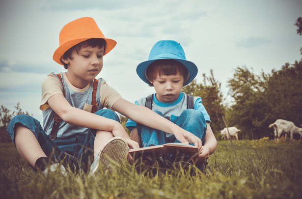 Two young boys sit on the grass, reading a book together during a family holiday. One wears an orange hat, the other a blue hat. Trees and goats dot the background beneath a partly cloudy sky.