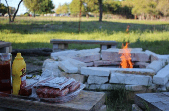 A fire pit with a burning fire is surrounded by benches in a grassy garden. In the foreground, hot dogs, ketchup, and mustard sit on a wooden table—perfect inspiration for your next DIY firepit ideas and outdoor barbeque.