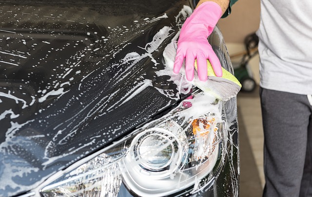 A person wearing a pink glove cleans the soapy headlamp and bonnet of a black car with a sponge, demonstrating great family vehicle upkeep.