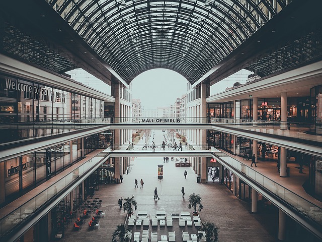 A spacious, modern shopping centre with multiple floors and a large arched glass ceiling reflects thoughtful urban planning. Shops line both sides and a sign reads "Mall of Berlin" above the central walkway.