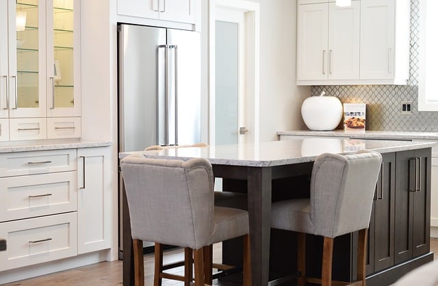A modern kitchen with white cupboards, a stainless steel fridge, a dark island with a grey worktop, and four upholstered bar stools. This stylish home improvement is sure to add value to your home. A large vase and framed picture sit on the counter.