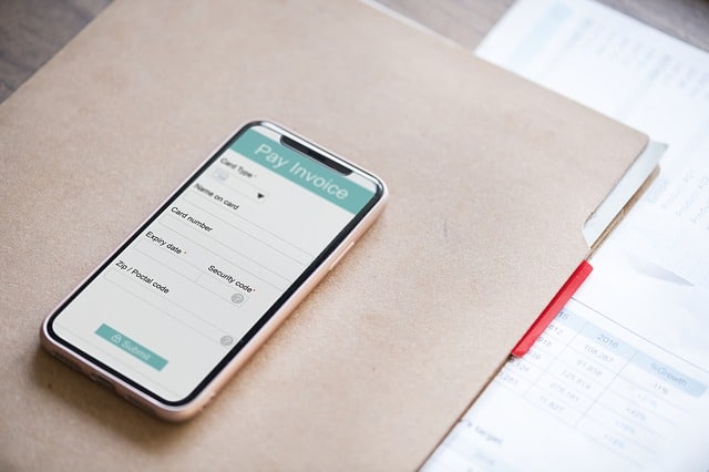 A smartphone displaying a "Pay Invoice" screen rests on a brown folder next to printed financial documents—an organised scene perfect for single income families focused on saving money and staying financially mindful.