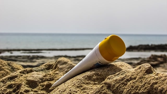 A tube of suncream with a yellow cap lies on sandy rocks at the beach, with the sea and hazy sky in the background—essential for a perfect day out.