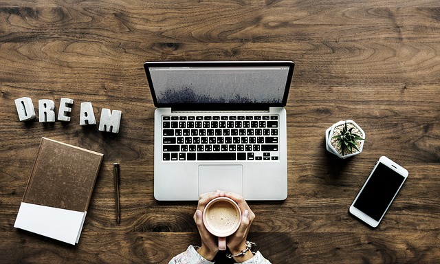 A person holding a cup of coffee sits at a wooden desk with a laptop, notebook, pen, mobile phone, small potted plant, and letter blocks spelling "DREAM," brainstorming CV tips to make your CV stand out.