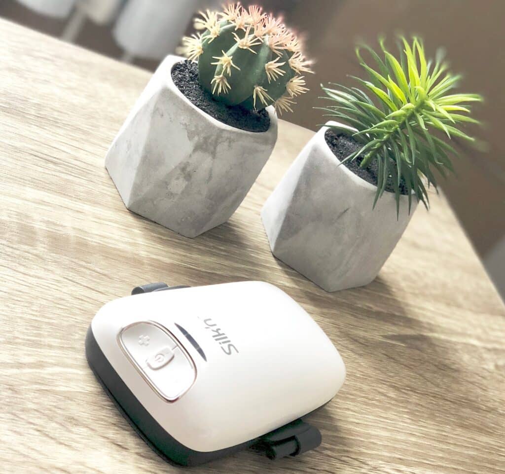 A white Silk'n Lipo device rests on a wooden surface beside two small potted plants, one cactus and one succulent, in geometric grey pots. Bright natural light highlights this serene Silk'n Lipo trial set-up.