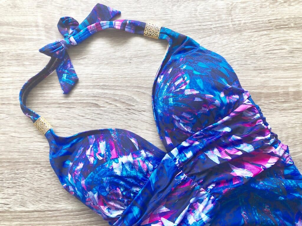 A close-up of a colourful Halter Plunge Swimsuit from Seaspray Ave, featuring blue, purple, and pink patterns. Gold accents on the straps and a tie at the neck add elegant details to this stunning piece lying on a wooden surface.