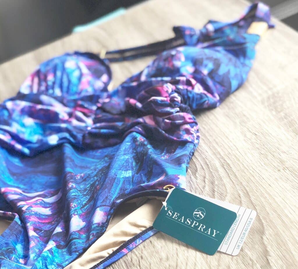 A blue and purple patterned halter plunge swimming costume lies on a wooden surface with a Seaspray Ave brand tag attached.
