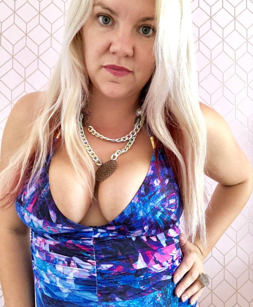 A woman with long blonde hair and light skin wears a blue and purple patterned halter swimsuit and layered necklaces, standing in front of a light pink geometric-patterned background, looking directly at the camera.