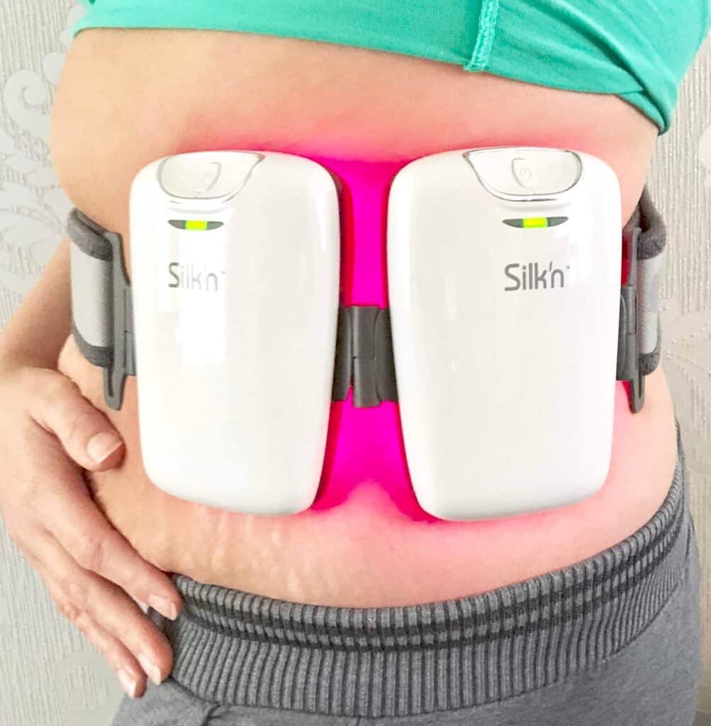 A person undergoing a Silk’n Lipo trial wears a device with two white panels emitting pink light on their abdomen, likely for a cosmetic or therapeutic treatment. The individual is dressed in a green top and grey trousers.