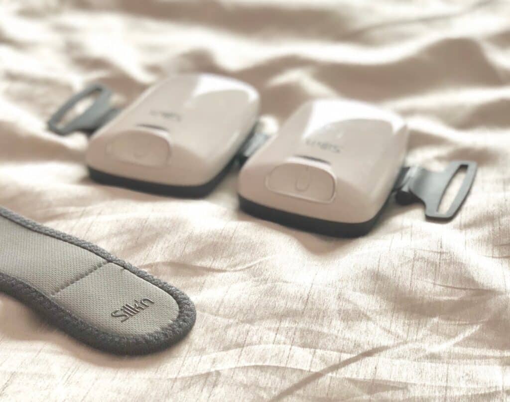 Two white electronic devices with buttons, labelled "Silk'n Lipo," rest on a soft, light-coloured fabric surface next to a grey, adjustable fabric strap—perfect for your Silk'n Lipo trial or update.