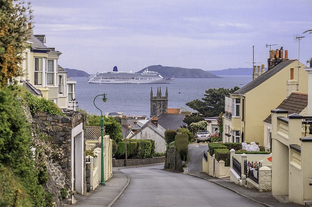 A gently sloping street lined with houses leads towards the sea, where a large white cruise ship sails past an island—a perfect scene for those seeking new ways to holiday or enjoy memorable UK holidays by the coast.