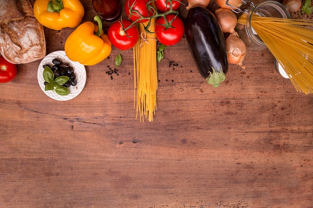 A wooden table with ingredients for Italian cooking—yellow and orange peppers, tomatoes, aubergine, onions, spaghetti, olives, basil, and a loaf of bread—arranged to inspire self-improvement and change your life through delicious meals.