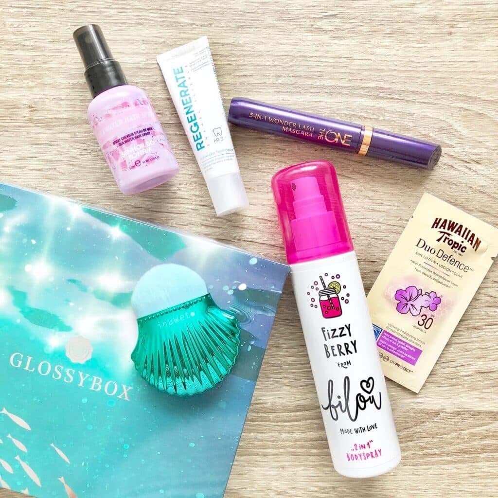 Assorted beauty products from a Glossybox beauty subscription, including a face mist, mascara, sunscreen sachet, GLOSSYBOX card, facial brush, hand cream, and “Fizzy Berry Bilou,” arranged on a light wood surface for an Under The Sea vibe.