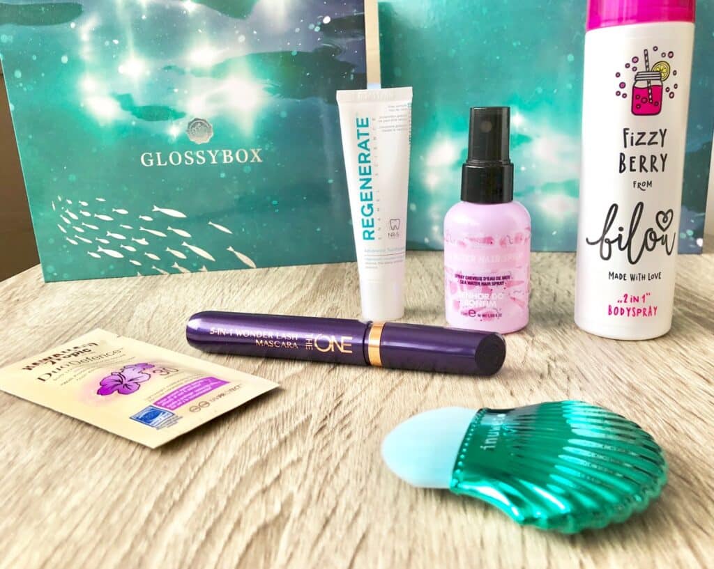 A selection of beauty and skincare products, including mascara, body spray, moisturiser, toothpaste, and a teal shell-shaped brush from the Glossybox "Under The Sea" beauty subscription, displayed on a wooden surface with the signature gift box in the background.
