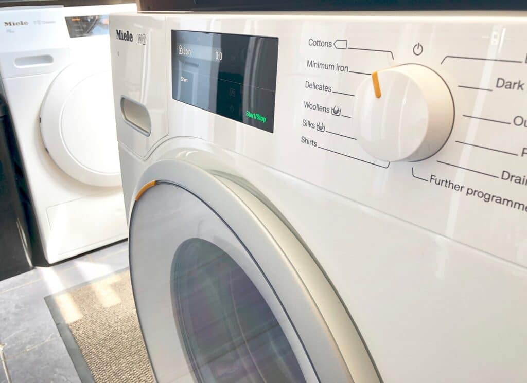 Close-up of a white washing machine with a digital display and programme dial showing Cottons, Delicates, Woollens, and Silks—ideal if you need to replace a broken appliance. Another appliance is visible in the background.