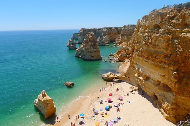A sunny beach with golden cliffs, turquoise sea, and scattered rock formations is one of the top holiday spots, where people—including new parents—relax under colourful umbrellas along the sandy shore.