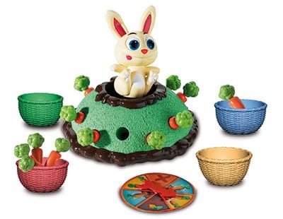 A colourful children’s game by University Games featuring a bunny on a green hill with fake carrots and green tokens, four coloured baskets, and a spinner. Duo up to win as the game pieces are arranged round the hill.