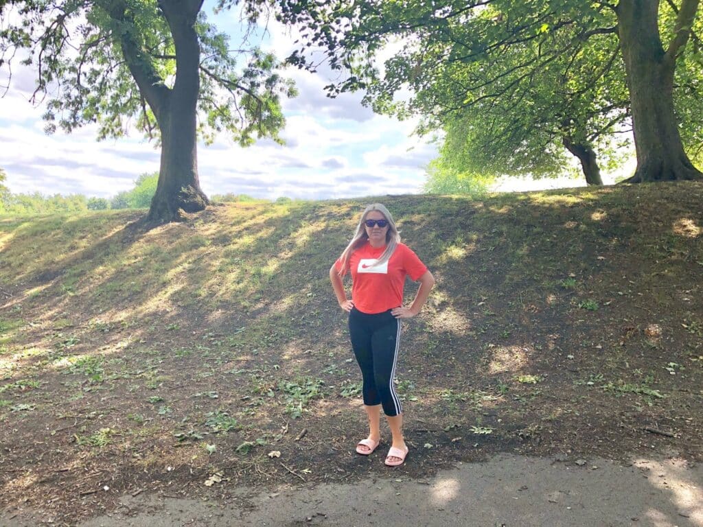 A woman in Mum Style, wearing a red shirt, black trousers from JD Sports, and sunglasses stands on a path in a sunlit park with trees and green grass under a partly cloudy sky.