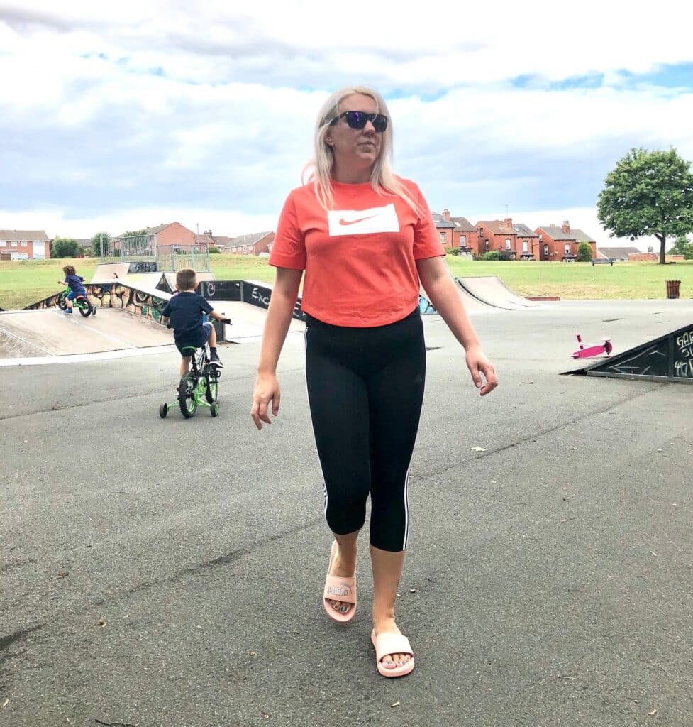 A woman in Mum Style wearing a red Nike shirt, black leggings, and sunglasses walks in a skate park. Children ride bikes and scooters nearby, with green grass, houses, and a tree under a cloudy sky. Perfect for the school run.
