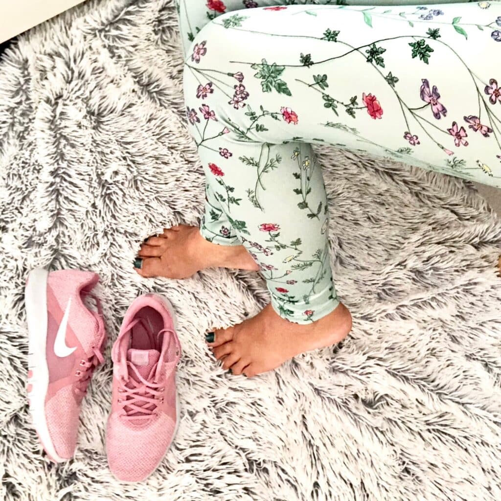 Person with floral leggings sits barefoot on a fuzzy grey rug next to a pair of pink Nike trainers—perfect Mum Style for the school run. Only legs and feet are visible, giving off those effortless JD Sports vibes.