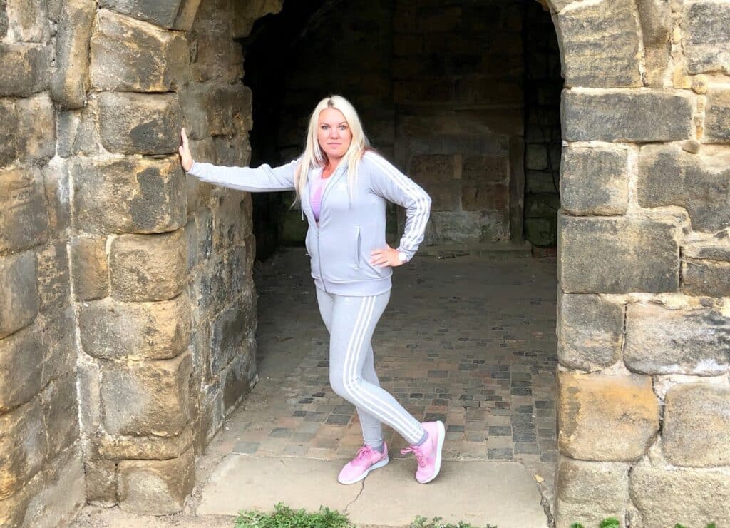 A woman in a light grey tracksuit and pink trainers channels Mum Style as she poses with one hand on the stone archway of an old brick structure, perfect for a stylish school run.