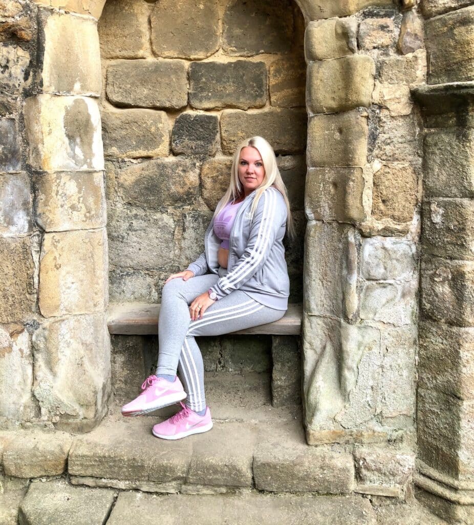 A woman in a grey tracksuit and pink trainers, perfect for School Run Mum Style, sits on a stone bench built into an old stone wall, posing for the camera with a relaxed expression.