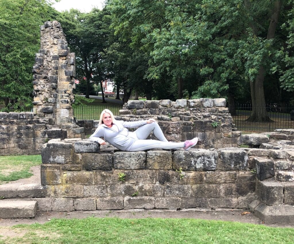 A woman in a grey tracksuit—perfect mum style for the school run—reclines on the stone ruins of an old wall, surrounded by green grass and trees in a park setting, embodying relaxed vibes and JD Sports flair.