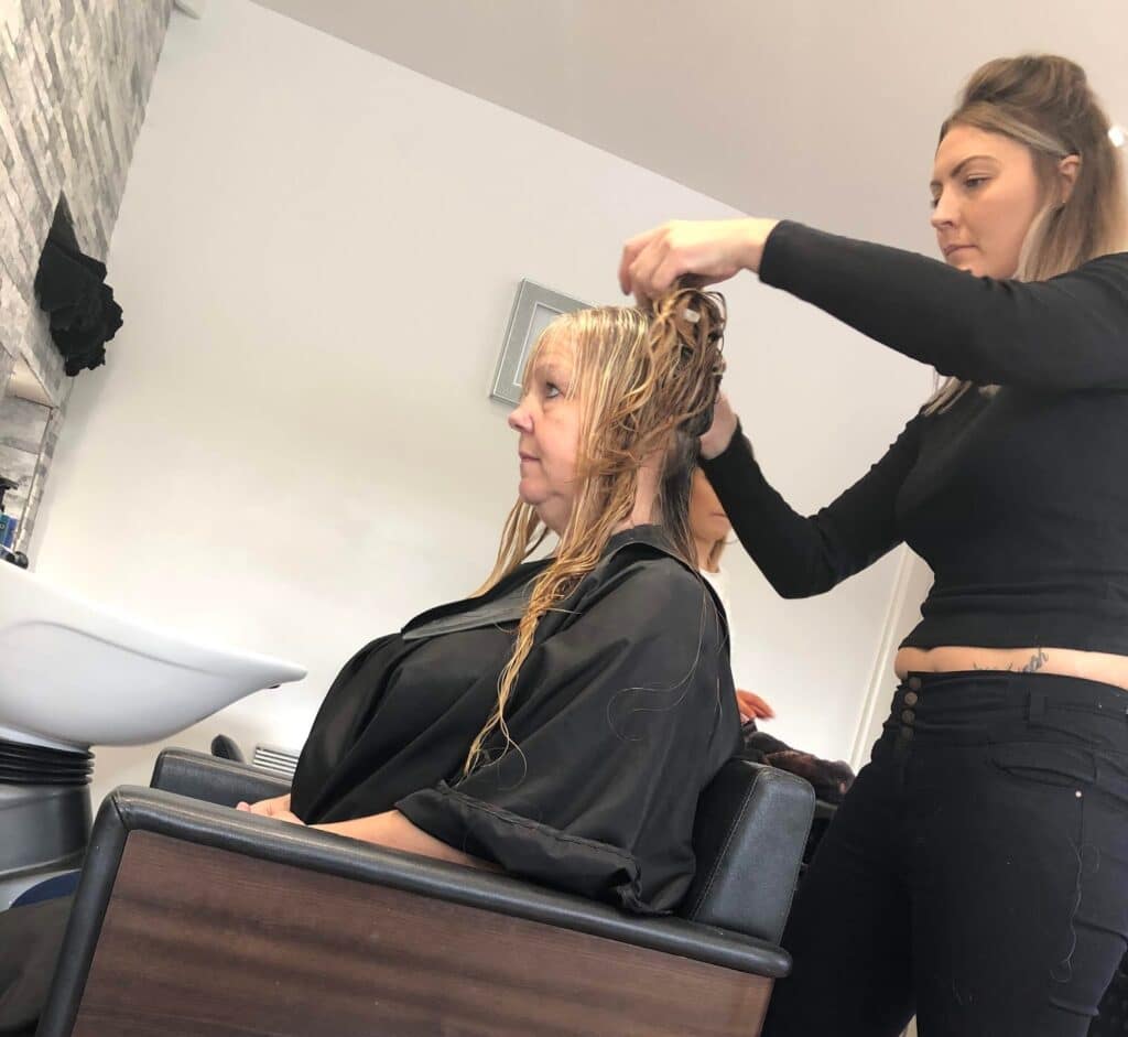 A woman sits in a salon chair with wet hair whilst a hairdresser stands beside her, both focused on the hair treatment—capturing the special moments of a Mum and Daughter Glam Day at a modern salon.
