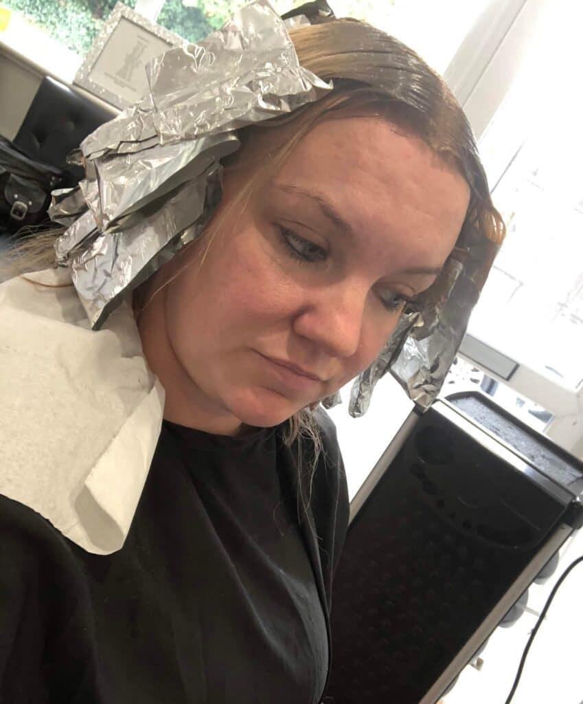A woman enjoys a Glam Day at the salon, sitting in a chair with sections of her hair wrapped in foil for highlights. She wears a black gown and white paper collar, looking down with a neutral expression—perfect for Mother Daughter Activities.