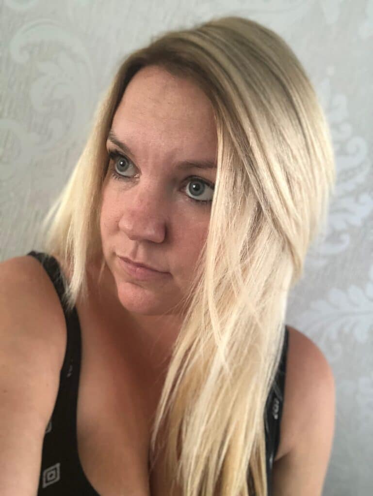 A woman with long, straight blonde hair and light eyes looks slightly to the side. Dressed in a black sleeveless top with a faint pattern, she captures the elegant spirit of a Glam Day—perfect for memorable Mother Daughter activities. The background features a pale, patterned wall.