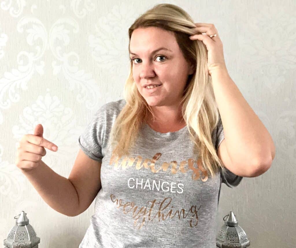 A woman with blonde hair wearing a grey “Kindness changes everything” t-shirt in gold and white lettering, pointing to the message on her shirt and touching her hair—perfect inspiration for a Mum And Daughter glam day, standing indoors against a patterned wall.