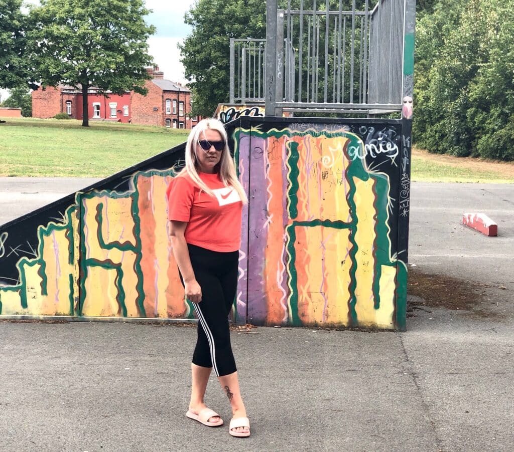 A woman in sunglasses, rocking Mum Style in a red Nike t-shirt from JD Sports, black leggings, and pink sliders stands in front of a colourful graffiti-covered ramp at an outdoor skate park on a cloudy day. Trees and a red brick building are behind her.