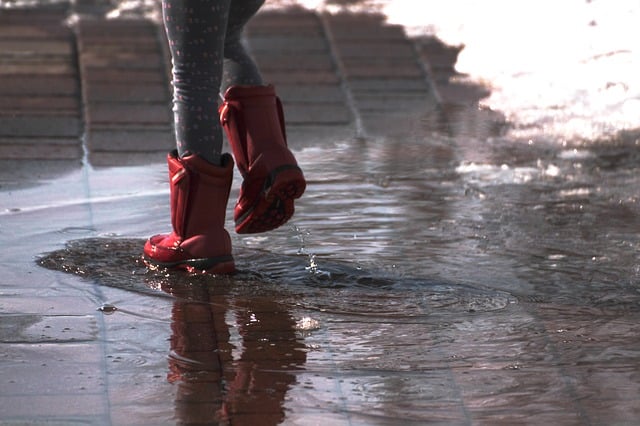 A child wearing red wellies and grey leggings walks through a puddle on a wet pavement with snow on the sides, enjoying messy play that supports healthy child development.