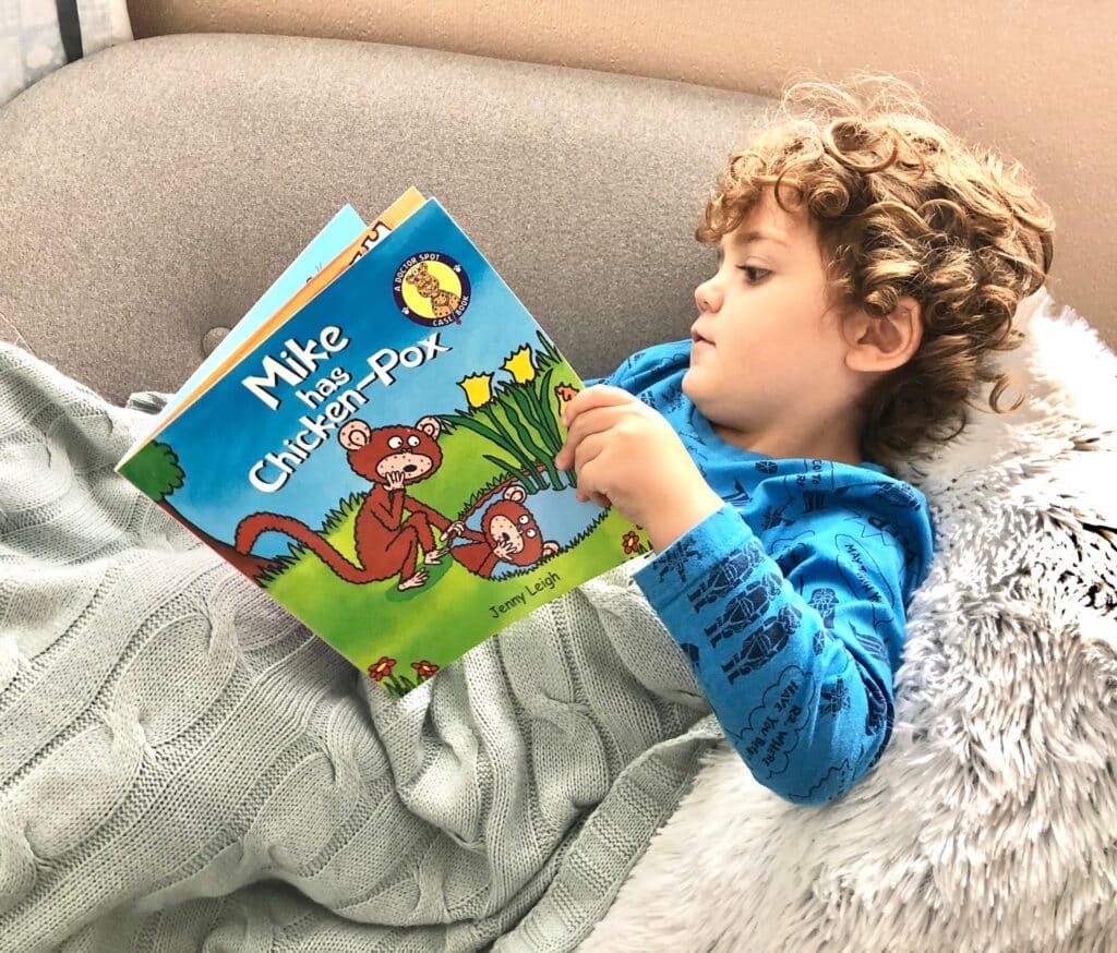 A young child with curly hair, wearing a blue shirt, lies on a grey sofa under a blanket and reads “Mike has Chicken Pox” by Jenny Leigh, exploring what to do and home remedies for chickenpox.