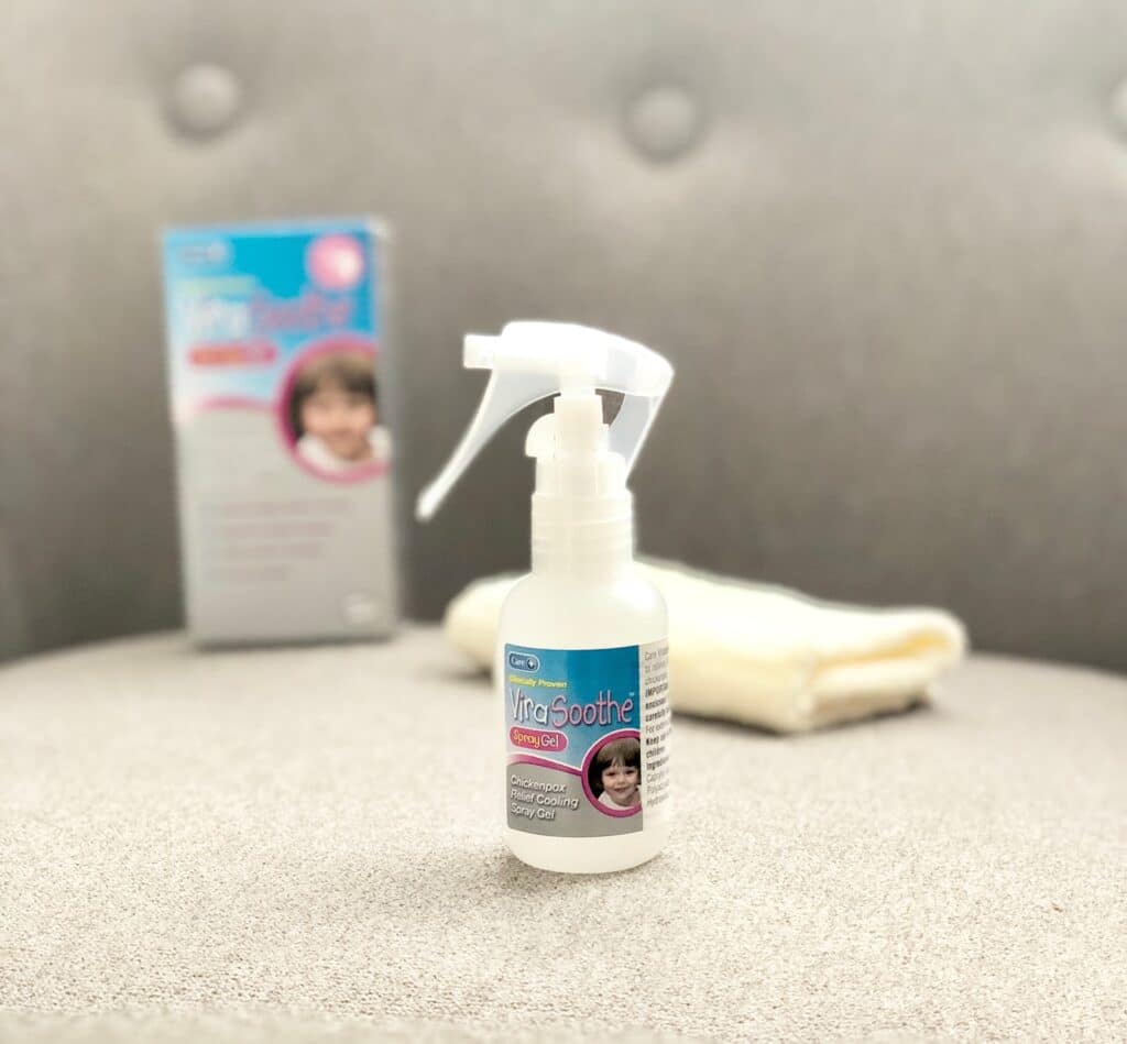 A white spray bottle labelled “ViraSoothe Spray Gel,” a gentle home remedy for chickenpox, sits on a grey cushion. The label features a smiling child’s face, while a blurred product box and folded cloth appear in the background.