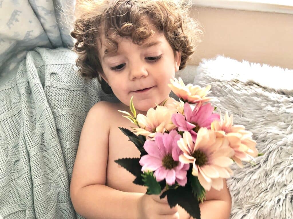 A young child with curly hair sits on a cosy seat, holding a bouquet of pink and cream flowers, looking at them with a gentle expression. Light filters in from a nearby window, creating a soft and warm home care atmosphere.