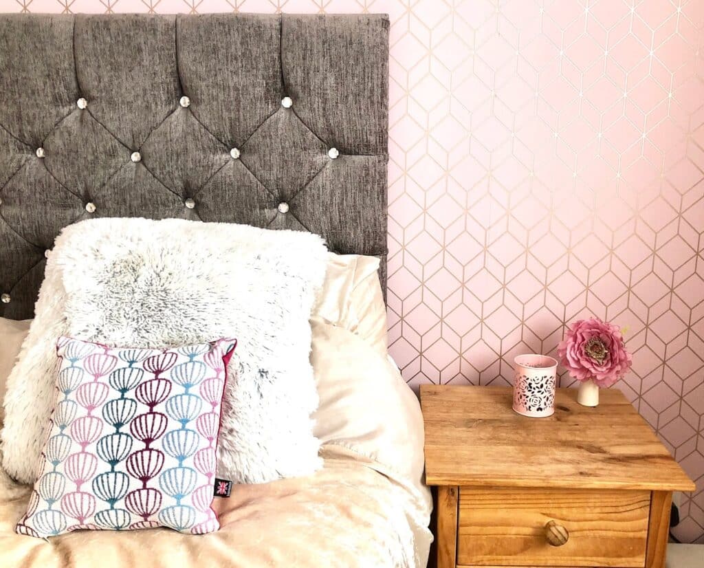 A cosy bedroom with a grey tufted headboard, fluffy white pillows, and a colourful cushion on a beige bed. The focal point is the wooden bedside table with a pink flower, pink mug, and candle against a pink patterned wall.