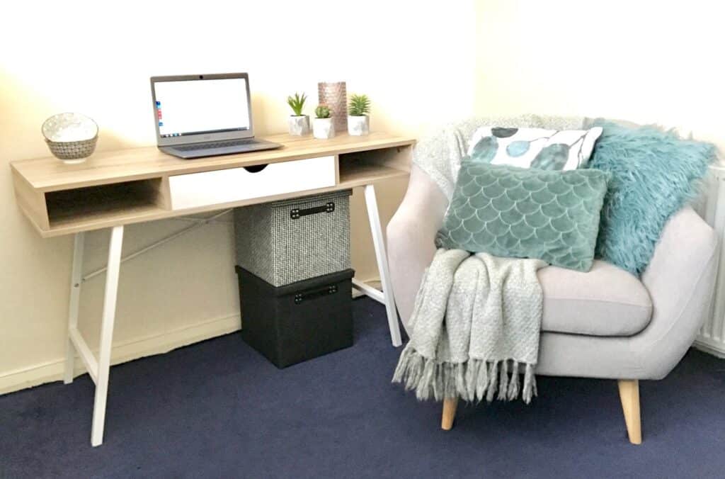 A cosy bedroom home office corner with a light wooden desk holding a laptop, decorative plants, and a candle, next to a cushioned armchair with pillows and a blanket—creating a serene focal point on the blue carpeted floor.