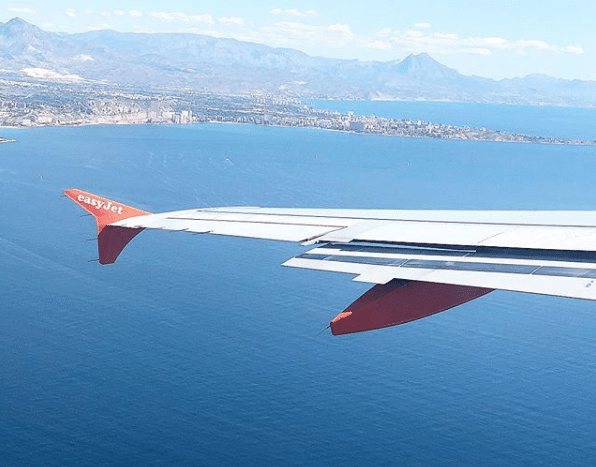 Aerial view from an easyJet aeroplane window showing the wing over a deep blue sea—perfect inspiration for holidaying abroad—with a coastal city and mountains in the distance under a clear sky.