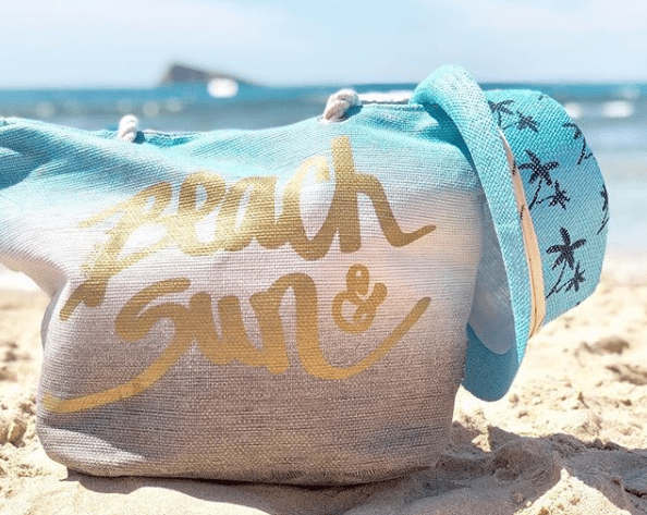 A blue and beige beach bag with "Beach & Sun" in gold sits on sandy beach beside a blue sunhat with starfish designs—perfect for holiday preparation or seeking travel tips whilst relaxing by the sea.