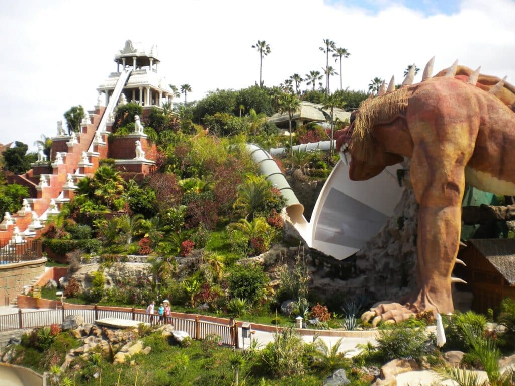 A lush, tropical water park in Tenerife features a large white slide descending from a temple-like structure and a tunnel slide emerging from the mouth of a giant dragon statue, with visitors walking along a nearby pavement.