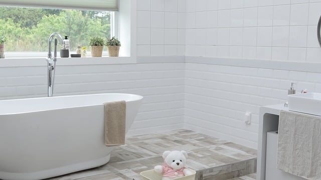 A bright, modern bathroom with a white freestanding bath, a beige towel draped over the edge, potted plants on the windowsill, and a white teddy bear in a basket—perfect inspiration for doing up our house with clever home improvements.