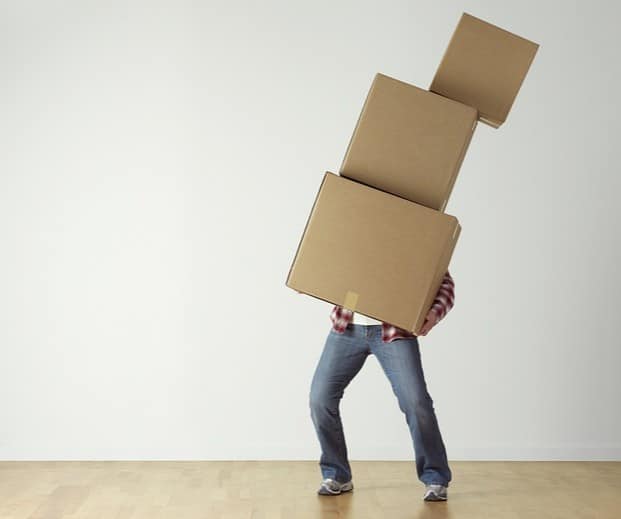 A person struggling to carry three large cardboard boxes stacked on top of each other, nearly losing balance in an empty room with a wooden floor and plain white wall—highlighting the need for stress-free moving.