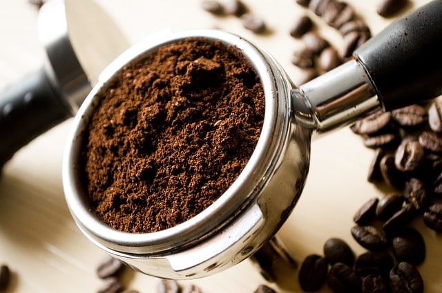 Close-up of a portafilter filled with freshly ground Fair Trade coffee, with whole coffee beans scattered around on a light surface.