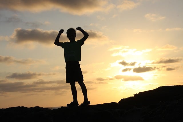 Silhouette of a child standing on rocks at sunset, arms raised in a triumphant pose, with a cloudy sky and the sun low on the horizon—capturing the spirit of fearless families across the UK embracing extreme adventure sports.